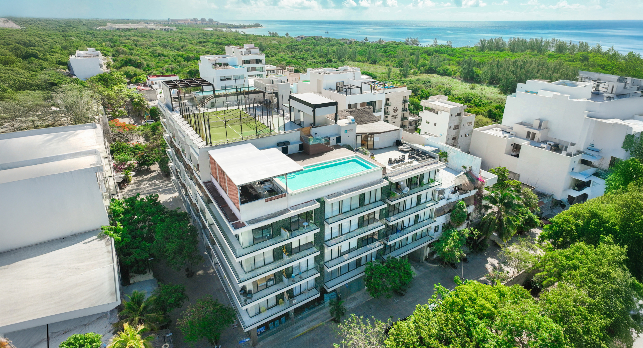 Vista aérea del edificio Macondo Quinta Avenida, un desarrollo con departamentos en venta en Playa del Carmen, que cuenta con alberca infinity en la azotea, cancha de pádel, áreas sociales y una espectacular vista al mar Caribe, rodeado de vegetación tropical y arquitectura moderna.