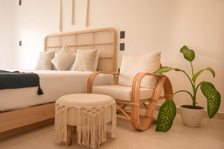 Habitación con cama matrimonial, cabecera de madera, sillón y banco de fibras naturales, planta decorativa en una maceta blanca y paredes en tonos claros con luz natural, dentro de un desarrollo con departamentos en venta en Playa del Carmen.