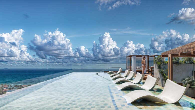 Alberca infinita en la terraza de Macondo Corazón con camastros sumergidos, vista panorámica al mar Caribe, pérgolas de madera, palmeras decorativas y cielo azul con nubes sobre Playa del Carmen. Un espacio exclusivo dentro de los departamentos en venta en Playa del Carmen que combina lujo y tranquilidad.