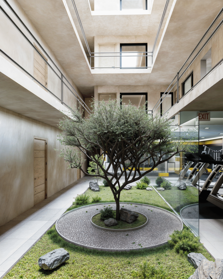 Solarium interior de Macondo Corazón, con un árbol central rodeado de jardín y piedras decorativas, pasillos con muros en tonos cálidos, barandales metálicos y ventanales que conectan con el gimnasio. Espacio diseñado para la contemplación y el bienestar dentro de los departamentos en venta en Playa del Carmen.