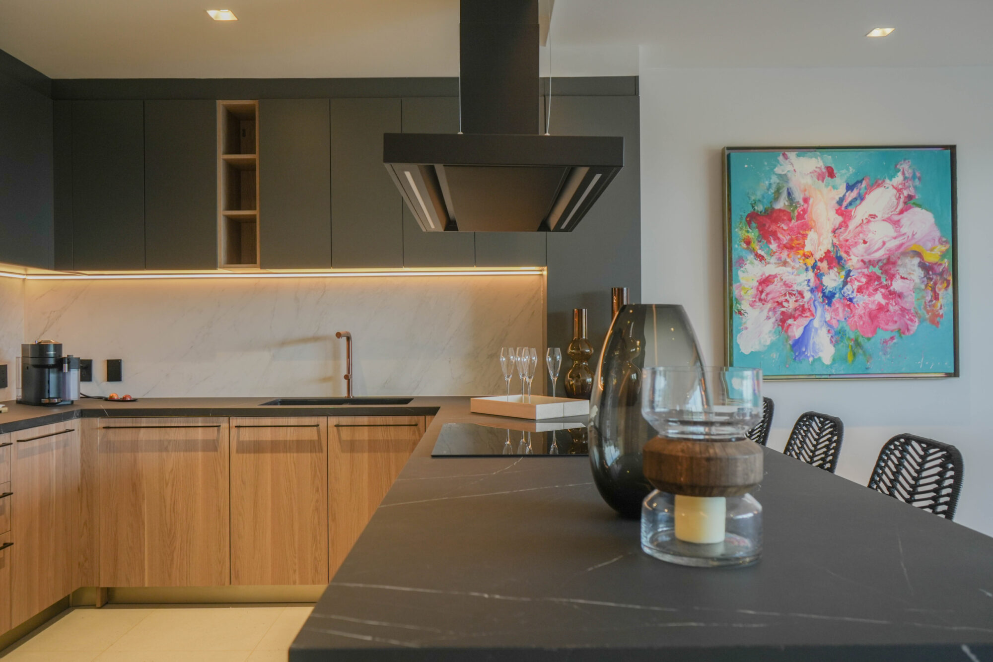 Cocina moderna en Macondo Playacar con acabados en madera natural, cubierta negra de mármol, gabinetes en tonos oscuros, iluminación cálida, campana de acero, grifería en color cobre y decoración con piezas de cristal y arte colorido. Un espacio distintivo dentro de los departamentos en venta en Playa del Carmen.