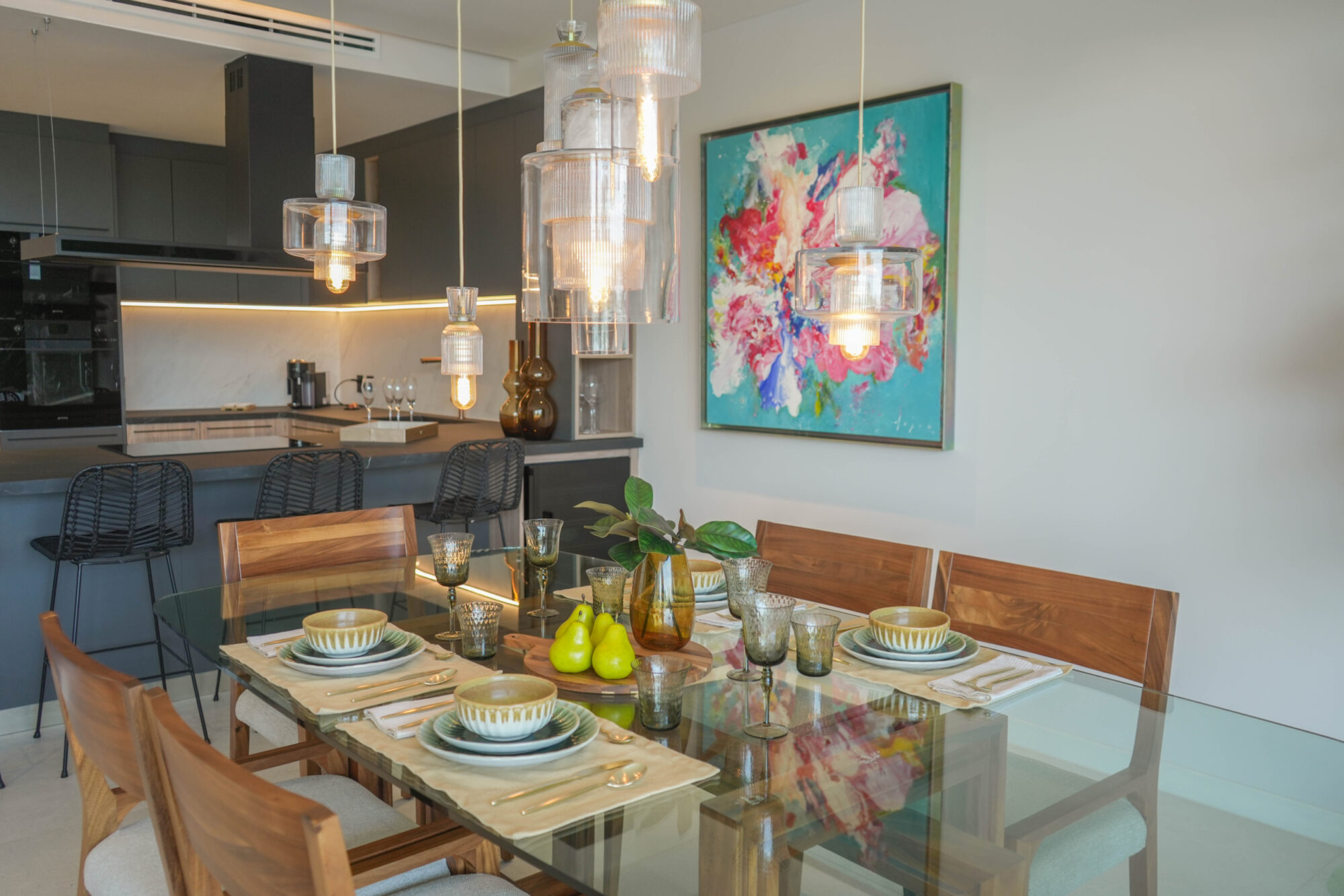 Comedor y cocina en una unidad de Macondo Playacar, con mesa de cristal y sillas de madera, vajilla artesanal, lámparas colgantes de vidrio, barra con sillas altas, gabinetes oscuros, arte colorido en la pared y detalles decorativos modernos. Espacio que refleja el estilo moderno de los departamentos en venta en Playa del Carmen dentro de este desarrollo residencial.