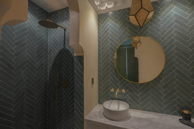 Baño en una unidad de Pink Riad Tulum con muros revestidos en azulejos verde oscuro, detalles dorados y un espejo circular enmarcado en oro. Inspirado en los hammams marroquíes, combina diseño contemporáneo con un aire exótico y sofisticado. Parte esencial de los departamentos en venta en Tulum.